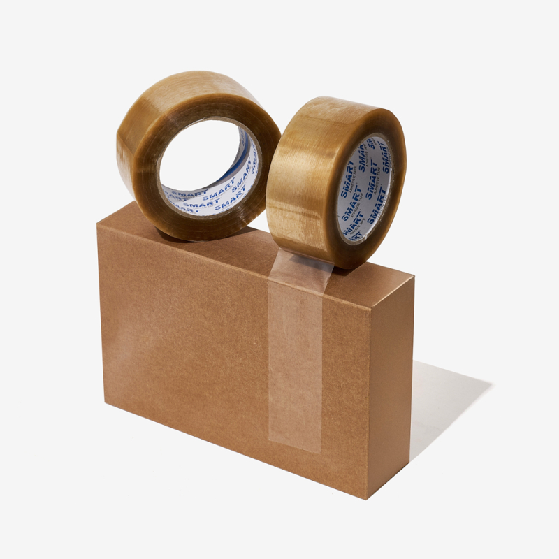 Zwei transparente, kautschukbasierte Klebebänder auf einem verschlossenen Versandkarton präsentiert.