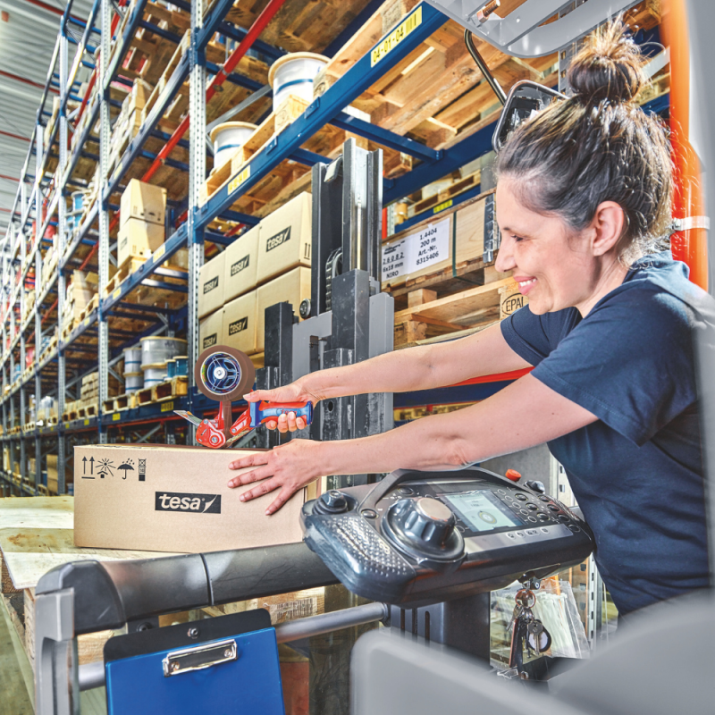Lagerarbeiterin verschließt einen Karton auf einer Palette mit braunem tesa® Klebeband