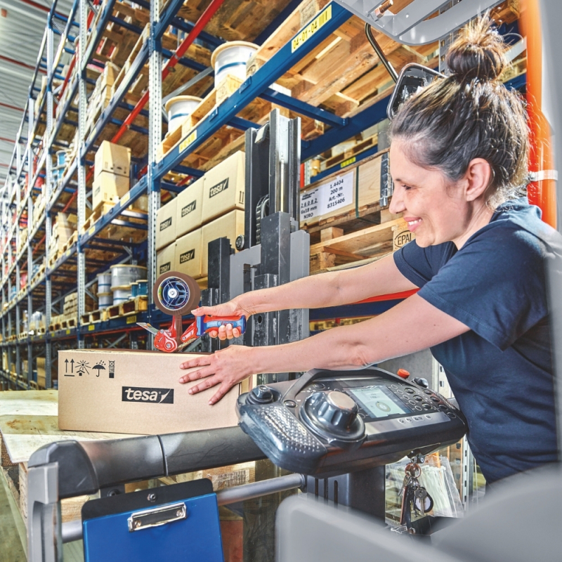 Eine Lagerarbeiterin verschließt einen Karton auf einer Palette mit braunem tesa® Klebeband.